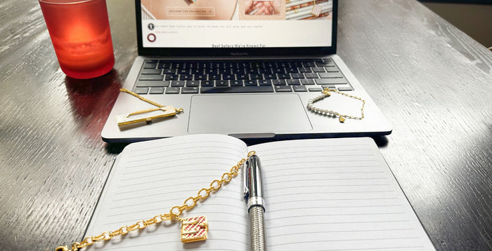 Secret Diary Book Bracelet (Magenta) laying on a notebook with a pen in front of a laptop with a Deco Fortune Locket (pearl) and a Fortune Cookie Pearl Bracelet